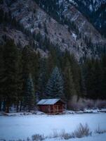 Secluded Cabin in a Snowy Mountain Landscape. A rustic red cabin sits peacefully amidst a snow-covered field, surrounded by towering evergreen trees and a dramatic mountain backdrop. photo