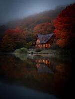 Illuminated Log Cabin Reflecting on Autumn Lake at Dusk. A stunning autumnal scene of a cozy cabin nestled amongst vibrant red and orange trees, its warm lights reflected on a tranquil lake. photo