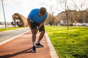 African American male runner experiencing knee pain on a city running track, frowning as he takes a break from exercising. photo