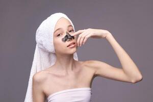 Young caucasian female applying charcoal face mask with towel on head for skincare routine. photo