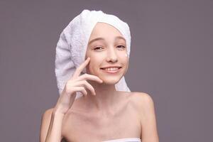 Young caucasian female skincare routine smiling with towel and cream on face against gray background. photo