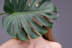 Mysterious young caucasian woman partially hidden behind large monstera leaf against gray background. photo