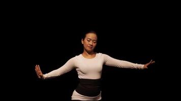 A dancer performing gracefully on stage, dressed in a white long-sleeve top and black pants. The background is dark, emphasizing the dancer's movements and expression. video
