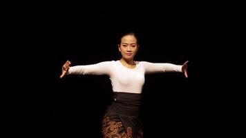 A dancer performing gracefully on stage, showcasing traditional movements. The dancer is dressed in a white long-sleeve top and a black skirt, with a focused expression, against a dark background. video