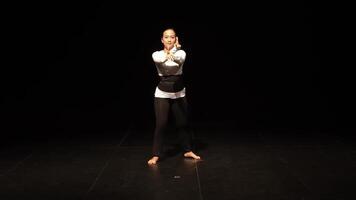 A dancer performing on stage in a dark setting, wearing a white long-sleeve top and black pants. The focus is on her graceful movement and posture, highlighting the art of dance. video