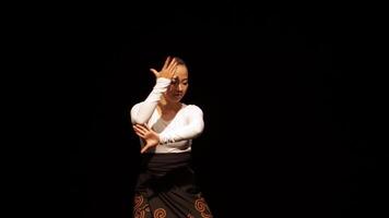 A dancer performing gracefully on stage, wearing a white long-sleeve top and traditional patterned skirt. The background is dark, highlighting the dancer's movements and expressions. video