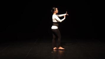 A dancer performing on stage with a black background, showcasing expressive movements. The dancer wears a white long-sleeve top and black leggings, emphasizing grace and fluidity in motion. video