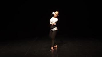 A dancer performing on stage in a dark setting, wearing a fitted white top and black pants. The focus is on the dancer's expressive movements and posture, highlighting grace and fluidity. video