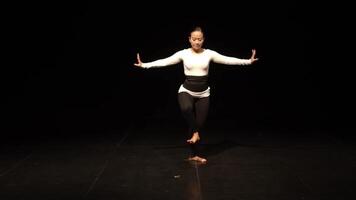 A dancer performing on stage with a black background, wearing a white long-sleeve top and black leggings. The dancer is in a graceful pose, showcasing movement and expression. video