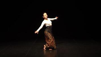 A dancer performing on stage in a dark setting, wearing a traditional outfit with intricate patterns. The dancer is in a graceful pose, showcasing fluid movements and cultural expression. video