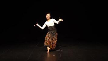 A dancer performing on stage, showcasing traditional attire with intricate patterns. The scene is illuminated against a dark background, highlighting the dancer's movements and expressions. video