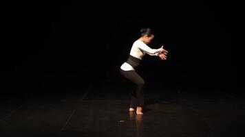 A dancer performing on stage with a black background, showcasing expressive hand movements and a graceful posture. The dancer wears a fitted white top and black leggings video