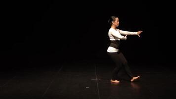 A dancer performing on stage in a spotlight, wearing a white long-sleeve top and black leggings. The background is dark, emphasizing the dancer's movements and expressions. video