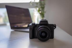 Modern digital camera with a prominent lens on a clean white desk, showcasing professional photography equipment and creative digital content creation tools with a laptop photo