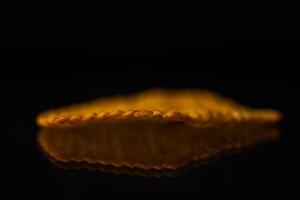 A single golden crispy snack chip displaying a distinct ridged texture, perfectly reflecting on a dark shiny surface, symbolizing indulgent pleasure of unhealthy food photo