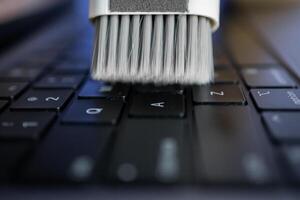White cleaning brush sweeps dust from a black laptop keyboard, representing essential technology maintenance for optimal productivity and device longevity photo