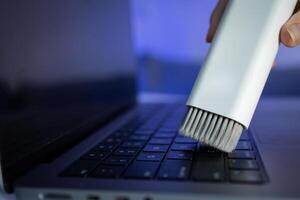 A white cleaning brush diligently sweeps dust off a sleek laptop keyboard, promoting essential electronic device care and regular maintenance for improved longevity photo