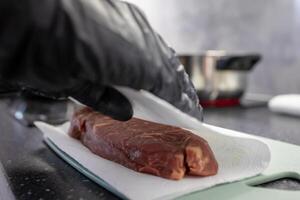 Professional chefs gloved hand gently patting dry raw beef steak with paper towel on cutting board for hygienic home kitchen food preparation photo