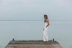 Asian woman in white outfit stands on wooden pier, calm lake, peaceful overcast, viewed from behind with contemplative mood, mindfulness, travel retreats, wellness photo