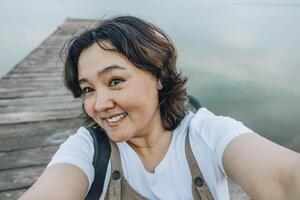 Asian woman smiling and looking at camera taking selfie on wooden pier with calm lake in background during travel adventure day trip concept of tourism vacation social media blog photo