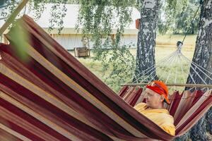 man relaxing in striped hammock under trees in red cap, yellow hoodie during summer camping retreat with glamping tents in background concept of outdoor leisure tourism resort marketing photo