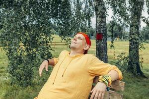 man in yellow hoodie and red cap resting with closed eyes on wooden bench under birch trees during warm summer day in rural outdoor area concept of vacation travel wellness retreat photo