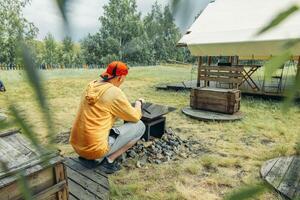 man in yellow squatting near outdoor fire pit surrounded by rocks with glamping tent in grassy field with trees, camping equipment, travel agency, outdoor adventure company, branches photo