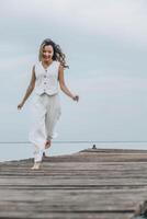 Asian woman running barefoot on wooden pier, calm sea horizon, dressed in white outfit under pale cloudy sky, mindfulness, wellness retreat, travel blog, motion blur, vertical photo