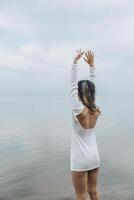 unrecognizable woman stands barefoot with hands up on wooden pier, hands up, calm sea horizon, in a white outfit, pale cloudy sky, mindfulness, wellness retreat, travel blog, vertical photo