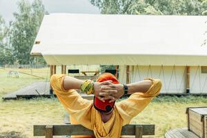 man in orange cap and yellow hoodie leaning back on wooden bench with hands behind head in front of glamping tent in forest meadow on summer day, travel eco tourism slow lifestyle retreat photo