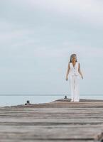 Asian woman in white outfit stands on wooden pier, calm lake, peaceful overcast, viewed from behind with contemplative mood, mindfulness, travel retreats, wellness, vertical, copy space photo