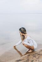 unrecognizable woman squatting barefoot at lake edge creating water ripples with fingers, gazing at water with serene expression, calm horizon under cloudy sky, mindfulness, wellness, retreat photo