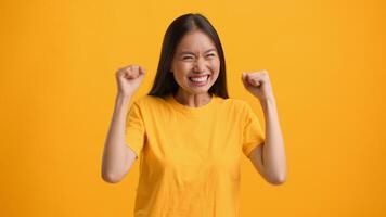 A millennial Asian woman expresses joy with a big smile and raised fists, showcasing her happiness. She stands against an isolated bright yellow background, embodying a lively lifestyle. video