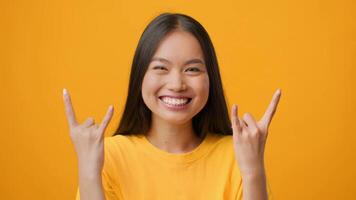 A millennial Asian woman is smiling widely while making a playful hand gesture. The bright yellow background enhances the joyful and lively atmosphere, perfect for lifestyle content. video