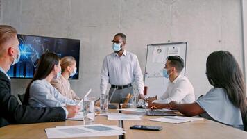 A group of six colleagues discusses business strategies in a contemporary office. Charts are displayed on a screen as ideas are shared. Masks are worn by all participants, promoting health safety. video