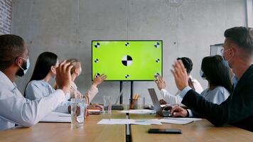 A diverse group of professionals in face masks is engaged in a collaborative session at a stylish office. They are attentively interacting with a large screen displaying visual content. video