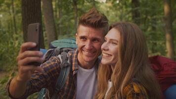 Two hikers share a joyful moment in the forest, capturing their adventure with a selfie amidst green trees and nature. Their backpacks hint at a day of exploration and fun outdoors. video