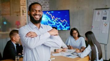 A focused business team collaborates in a contemporary office setting. The leader African American man stands confidently in front, while colleagues discuss strategies and project data video