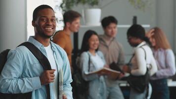 A group of diverse college students gathers in a contemporary study area, engaging in discussions and sharing ideas. One student African American guy smiles in the foreground, holding notebooks. video
