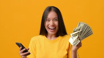 A joyful woman holds a bundle of cash in one hand and a smartphone in the other against a vibrant yellow background. She appears thrilled, capturing a moment of achievement and celebration. video