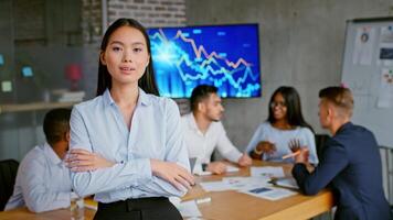 A professional Asian woman stands confidently in a stylish office, while a team discusses data trends in the background. The atmosphere reflects determination and teamwork video