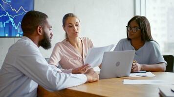 Tres multiétnico profesionales son comprometido en un enfocado discusión alrededor un ordenador portátil en un moderno oficina. ellos son analizando documentos y estrategia para negocio éxito durante tiempo de día horas. video