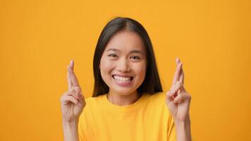 A millennial Asian woman with long hair smiles broadly while crossing her fingers in a cheerful gesture. The bright yellow background enhances her vibrant expression, radiating positivity. video