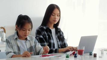 In a bright kitchen, a mother and her daughter engage in a creative activity, painting together. They share a moment of joy and affection while surrounded by art supplies and a laptop. video
