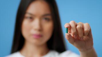 A millennial Asian woman with long dark hair showcases a small green capsule in her hand. She stands against a vibrant blue background, emphasizing her expression of curiosity and engagement. video
