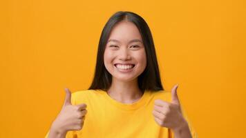 A young Asian woman smiles brightly while giving two thumbs up against a vibrant orange background. Her joyful expression radiates positivity and confidence, embodying a cheerful lifestyle. video