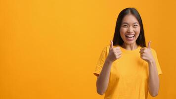 A young Asian woman with long hair smiles enthusiastically while pointing both hands to the sides. She wears a vibrant yellow t-shirt and stands against a plain yellow background, conveying joy. video