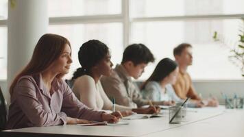 Diverse group of college students work diligently at a long table in a modern classroom. They are writing notes and collaborating on projects amid a bright environment. video