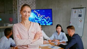 A confident woman stands in the foreground, showcasing leadership while her colleagues collaborate around a table. Behind them, a digital screen displays fluctuating graphs, emphasizing success. video