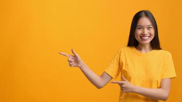 A joyful Asian woman wearing a yellow shirt is posed against a bright yellow background. She is smiling and pointing with enthusiasm at copy space, exuding a cheerful lifestyle vibe. video